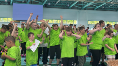 Viele Kinder in leuchtend hellgrünen T-Shirts stehen in einer Gruppe vor den Fenstern eine Sporthalle zusammen. Sie lachen, winken und tanzen.